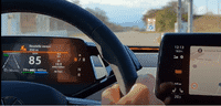 A hand gripping a car steering wheel with a view of the windshield and rear-view mirror.