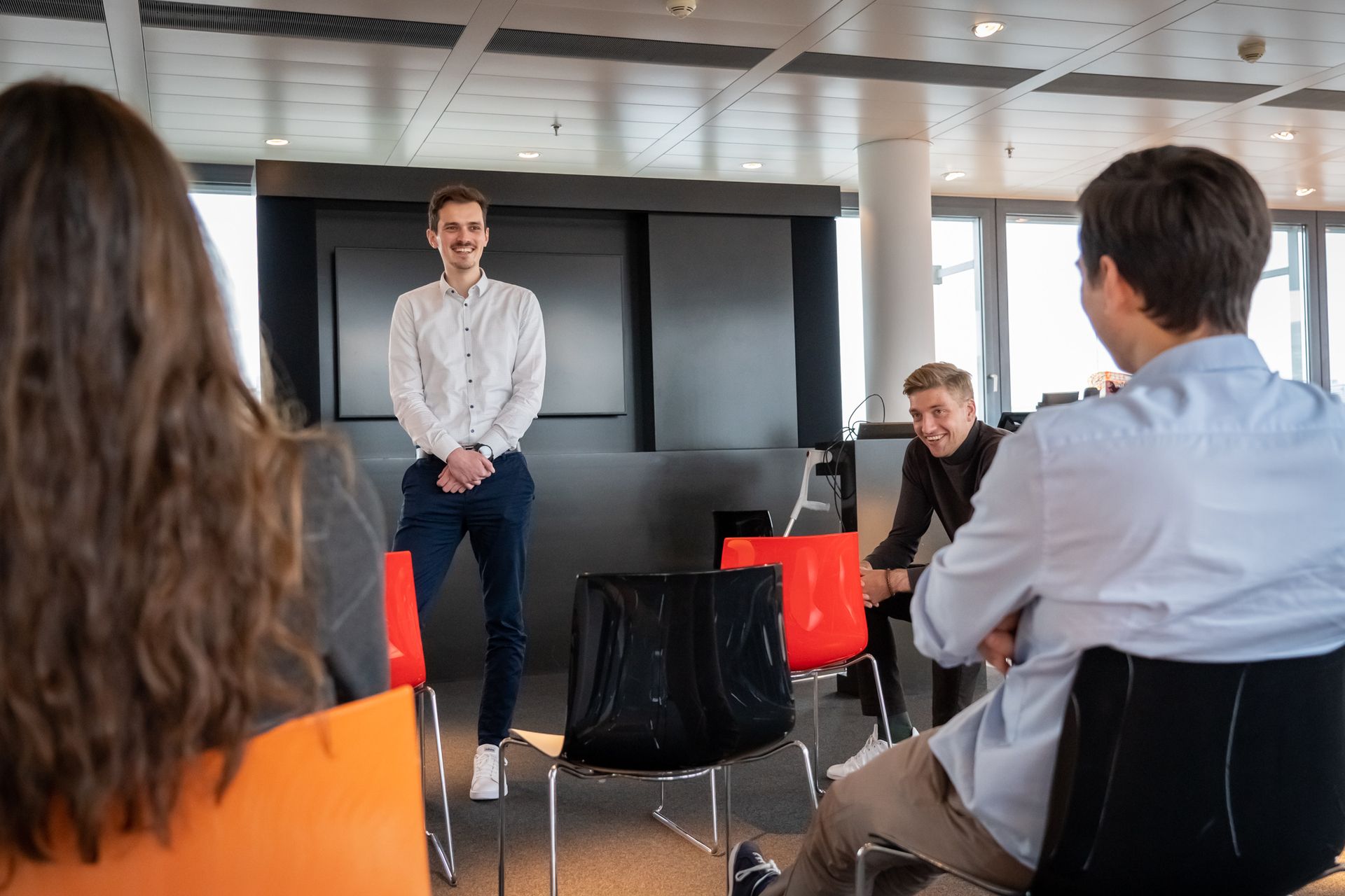 Eine Gruppe von Menschen sitzt auf Stühlen, während ein Mann vor einer schwarzen Wand steht.
